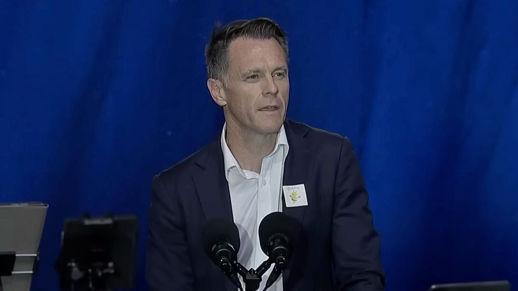 Man in a dark suit speaking at a podium with two microphones, blue backdrop and a small sticker on his lapel.
