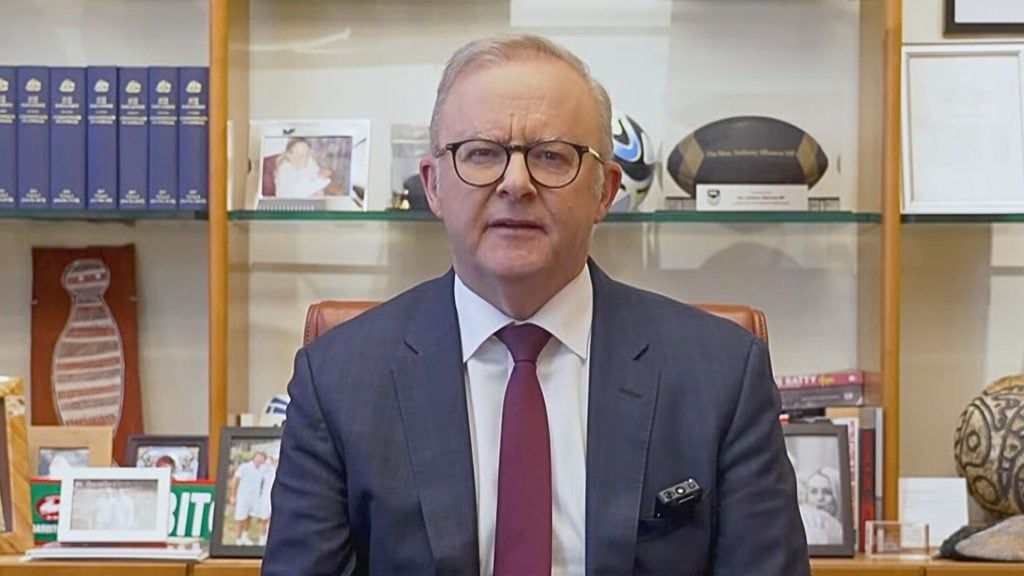 Albanese with short gray hair and round glasses wearing a dark suit and burgundy tie sits facing the camera in an office, with glass shelves behind him holding framed photos, blue binders and sports memorabilia including a soccer ball and rugby ball.