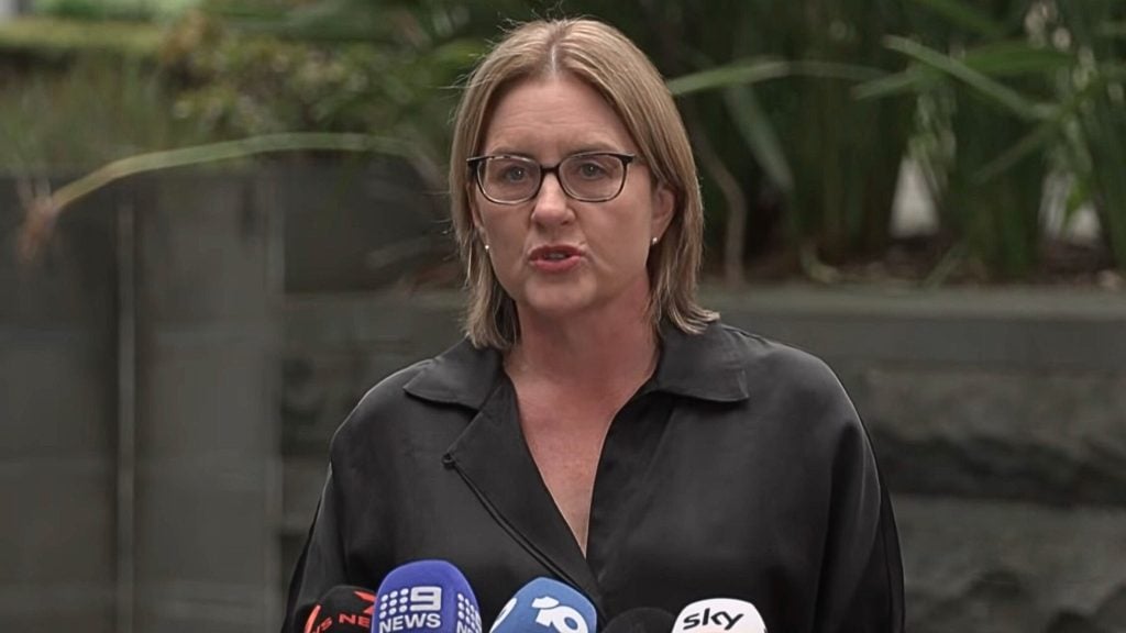 Allan in glasses speaking at an outdoor press conference with multiple TV microphones.