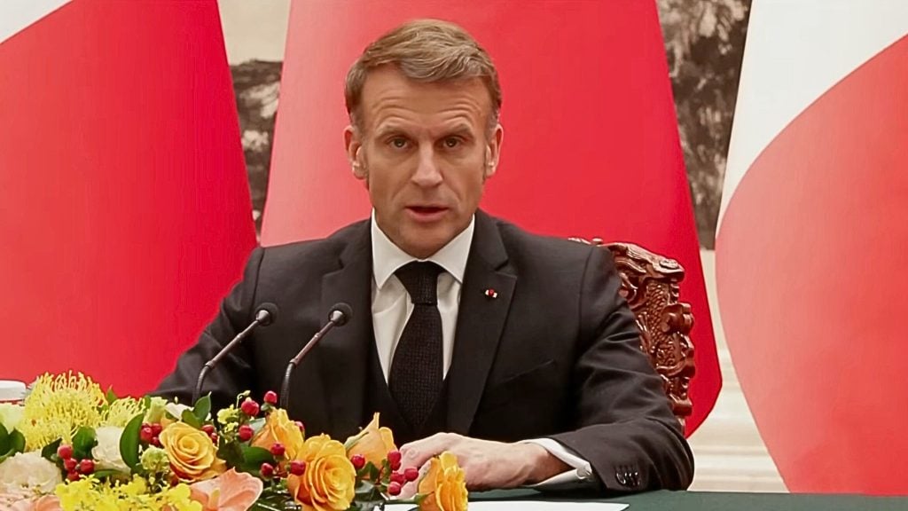 A middle-aged man with light brown hair in a dark suit and black tie sits at a table speaking into two microphones, a bright arrangement of yellow and orange flowers with red berries in front of him and large red flags or banners and an ornate carved chair behind, his expression serious.