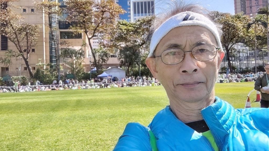Wong Kwok-ngon wearing glasses, a white sweatband, and a blue jacket takes a selfie on a sunny grassy park with a crowd, trees, and tall buildings in the background.