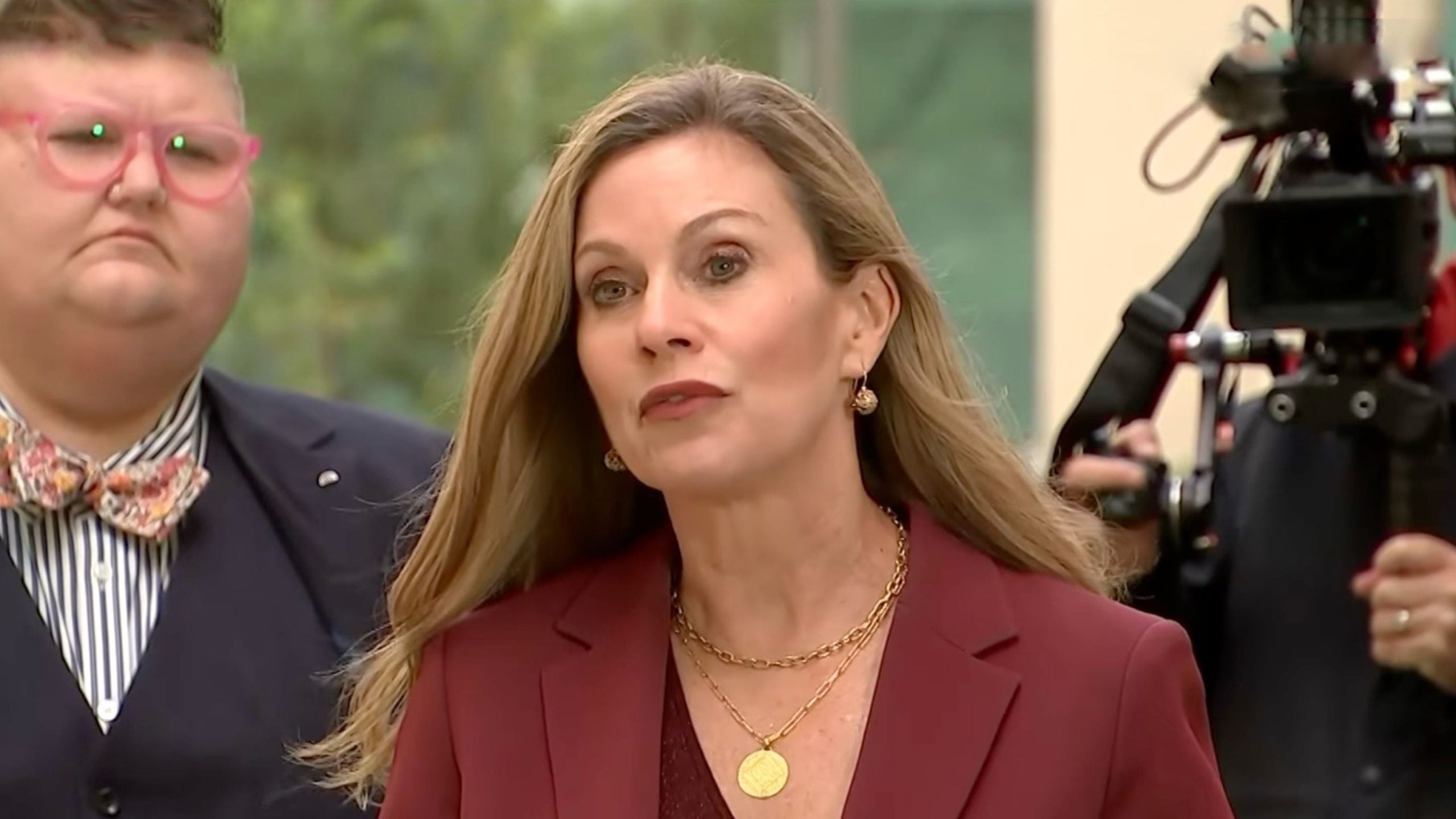 Inman Grant in a burgundy blazer and layered gold necklaces speaking to cameras, with a person wearing pink glasses and a floral bow tie standing behind her.