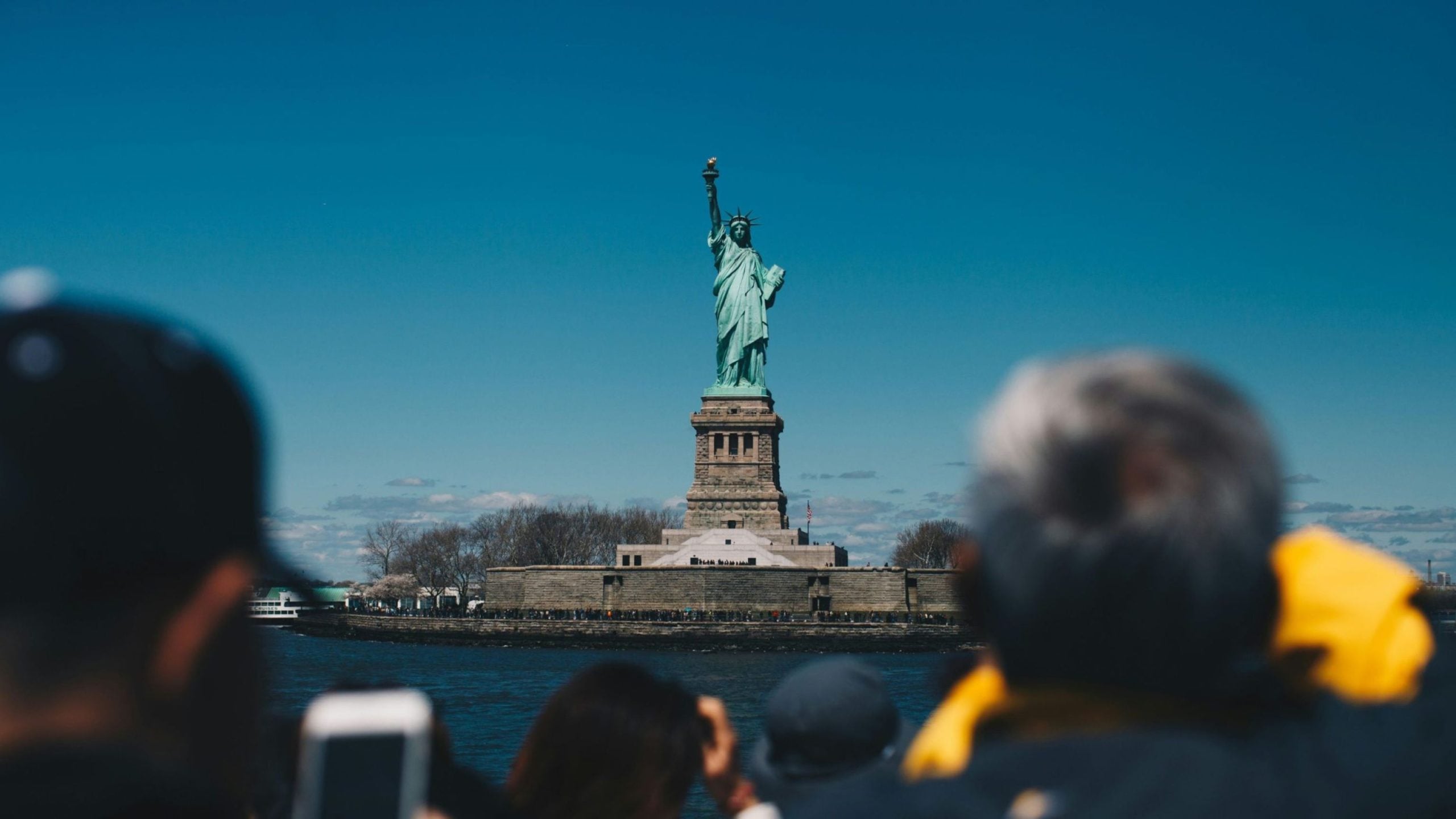 Statue of Liberty standing on its pedestal on Liberty Island under a clear blue sky, framed by blurred boat passengers in the foreground.