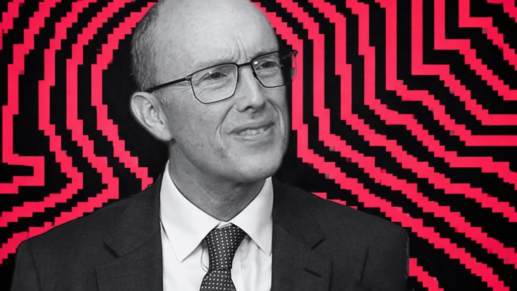 Jonathan Hall in a suit and glasses smiling against a bold red-and-black zigzag patterned background.