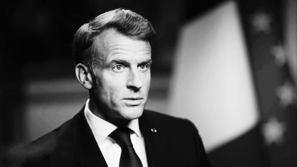 Close-up black-and-white portrait of Macron in a suit and tie with a blurred flag in the background.
