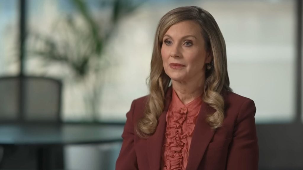 Inman Grant with curled blonde hair wearing a maroon blazer and ruffled coral blouse, sitting in a softly lit office with a blurred plant behind her.