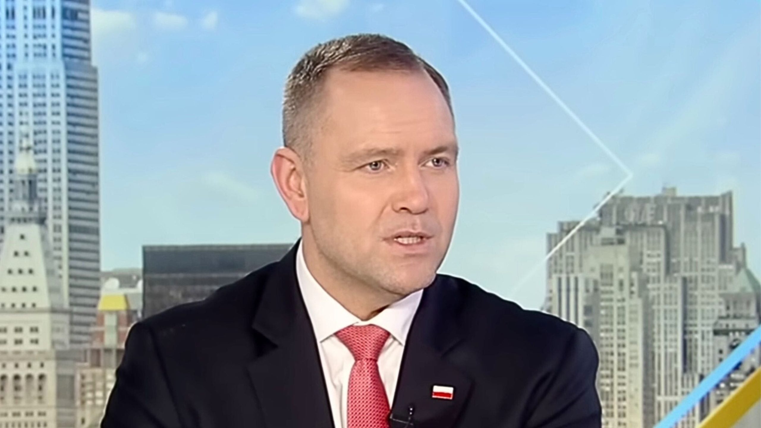 President Karol Nawrocki in a dark suit and red tie with short-cropped hair and a small red-and-white flag lapel pin speaking against a city skyline
