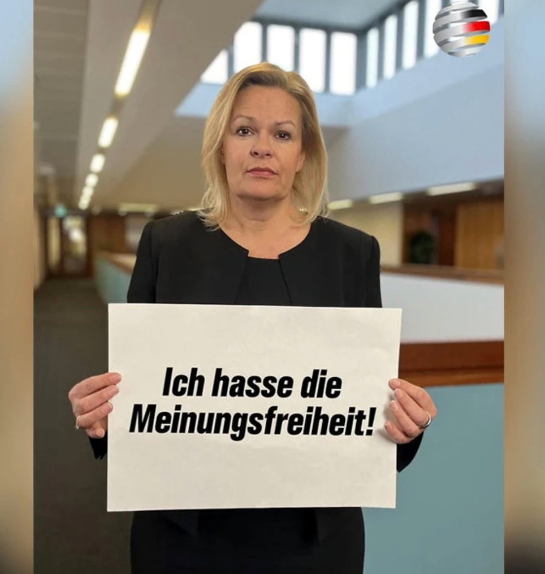 Faeser in a black jacket standing in an office corridor holding a sign reading "Ich hasse die Meinungsfreiheit!"