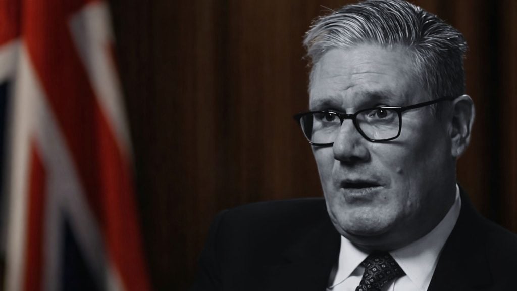 Starmer in glasses and suit speaking at an interview, close-up with a blurred Union Jack flag in the background.
