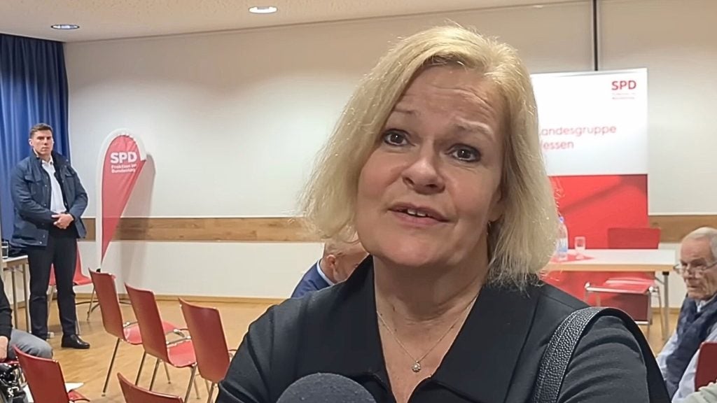 Faeser speaking to camera in a meeting room with red SPD banners and rows of empty chairs behind her.