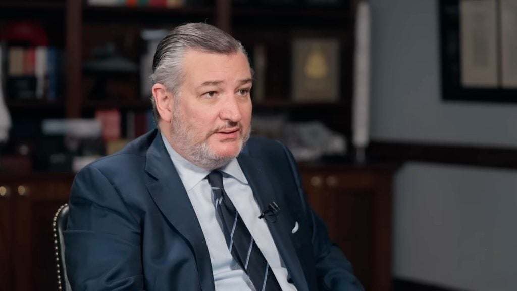 Cruz with slicked-back hair and beard wearing a dark suit and striped tie, seated in an office with bookshelves and speaking.