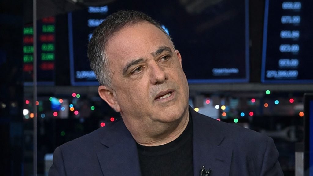 Kramer in a dark blazer and black shirt speaking in a studio with blurred monitors and colorful lights behind him