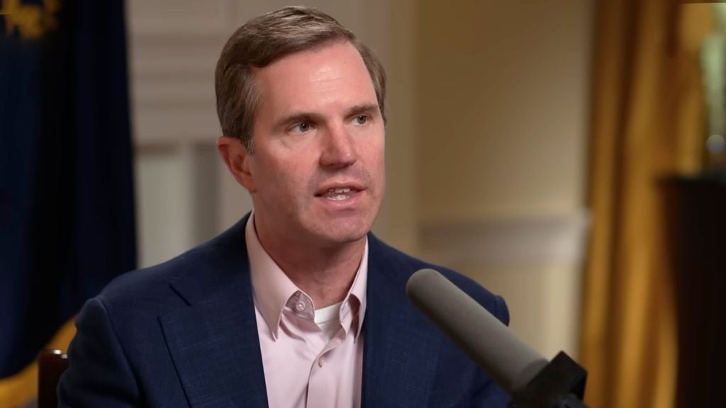 Governor Andy Beshear in a pink shirt speaking into a large microphone during an indoor interview.