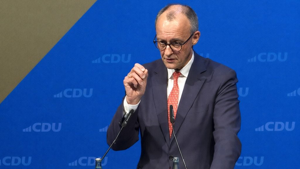 Merz wearing glasses, a dark suit and red tie speaking at a podium with a blue CDU backdrop, gesturing.