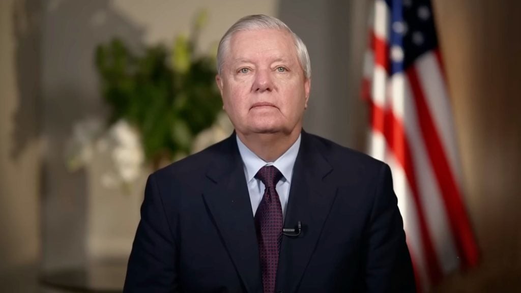 Graham with silver hair in dark suit and patterned tie, wearing a lapel microphone, US flag blurred in background