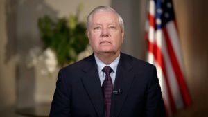 Graham with silver hair in dark suit and patterned tie, wearing a lapel microphone, US flag blurred in background