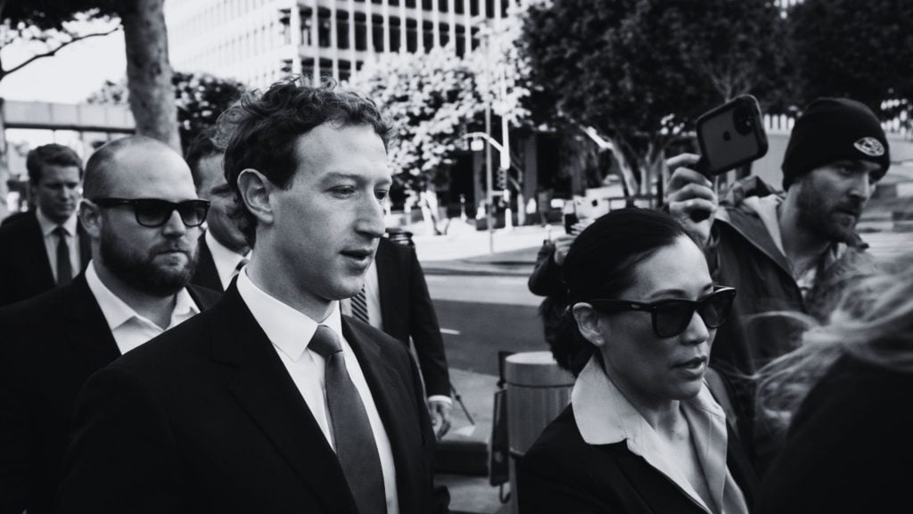 Black-and-white photo of a suited Zuckerberg with short curly hair walking with security as someone films on a phone.