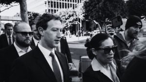 Black-and-white photo of a suited Zuckerberg with short curly hair walking with security as someone films on a phone.