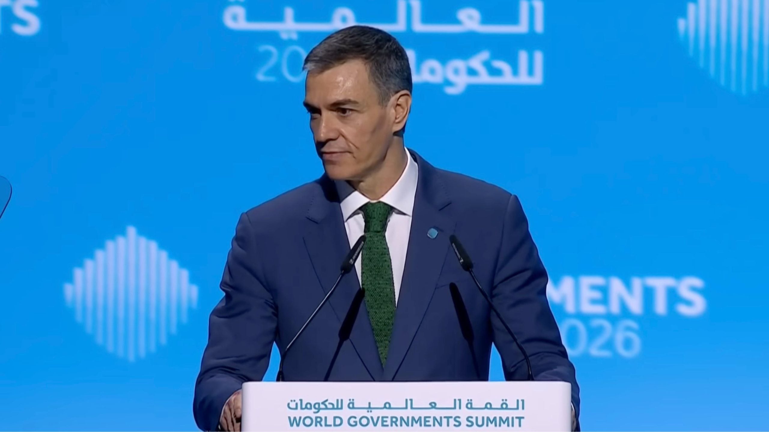 Pedro Sánchez in a dark suit and green tie speaking at a podium labeled World Governments Summit against a blue backdrop.