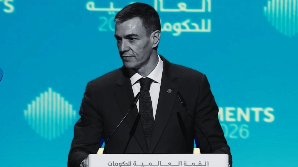 Pedro Sánchez in a dark suit and green tie speaking at a podium labeled World Governments Summit against a blue backdrop.