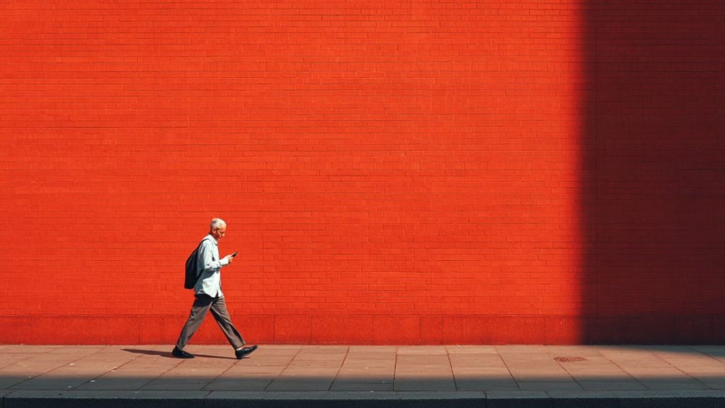 Elderly man walking along a sidewalk in front of a bright red brick wall, looking at his phone and wearing a backpack.