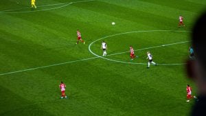 Soccer aerial view of white and red-striped players near center circle, ball in air and blurred spectator head at right.