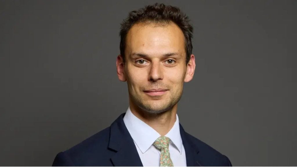 Simons in a navy suit, white shirt and pale green patterned tie, smiling at the camera against a plain gray background.