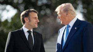 Macron and Trump in suits facing each other outdoors, one in black with a dark tie, the other in navy with a blue tie and a U.S. flag lapel pin.
