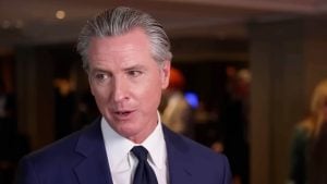 Middle-aged man with slicked-back gray hair in a blue suit, white shirt and dark tie speaking at an indoor event with a blurred crowd behind him.