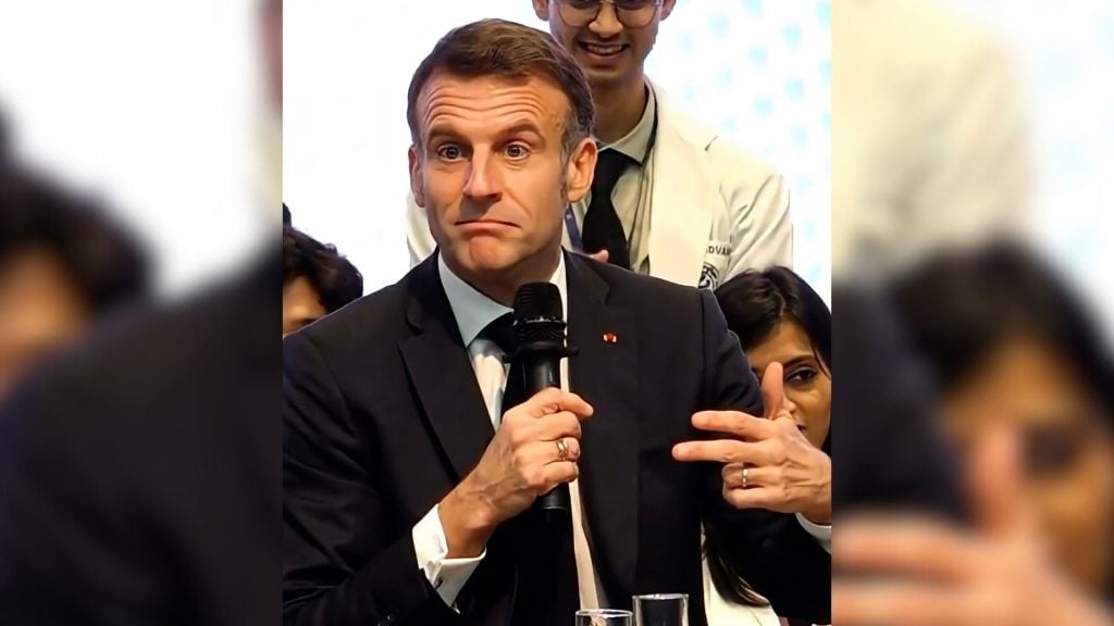 Macron in dark suit and tie holding a microphone and gesturing with his hand, looking slightly surprised with people behind him.