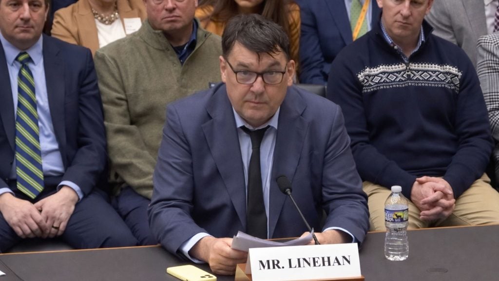 Linehan seated at a hearing table, holding papers in front of a nameplate and microphone.