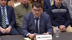 Linehan seated at a hearing table, holding papers in front of a nameplate and microphone.