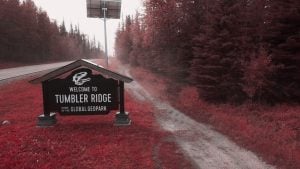 Welcome to Tumbler Ridge sign with dinosaur skull logo beside a roadside gravel path and red-tinted forest