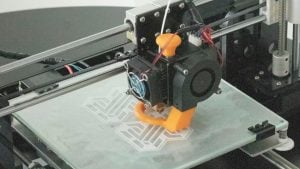 Desktop 3D printer extruder with black fan and orange knob printing an orange curved part on a glass print bed with white lattice infill.