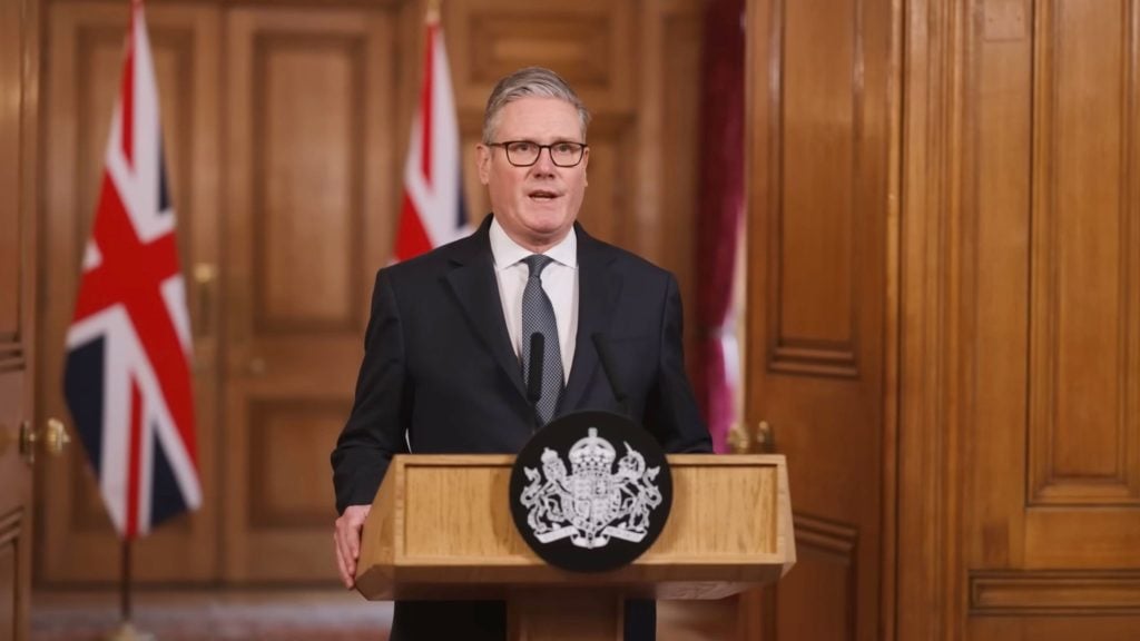 Starmer in suit and glasses standing at a wood podium with a government crest, British flags and wood-paneled room behind him.