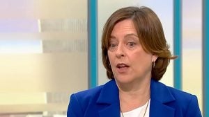 Dawes with short brown hair and gold hoop earrings speaking, wearing a blue blazer and white top against a pale studio background