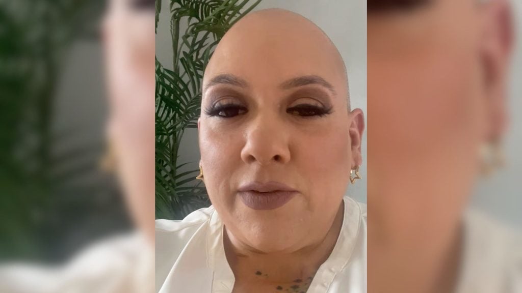 Close-up of a bald person with dramatic eye makeup, mauve lipstick, star-shaped gold earrings, and a leafy plant in the background.