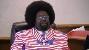Afroman wearing a red, white, and blue American flag–patterned suit and matching star-spangled tie and sunglasses, seated indoors.