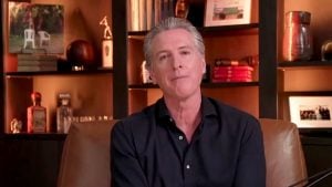 Newsom with gray hair in a dark shirt seated in a chair before lit shelves of books, trophies and photos.