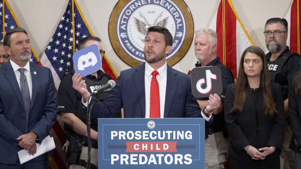 Uthmeier in suit at podium holding Discord and TikTok logos with sign reading "Prosecuting Child Predators," officials behind him.