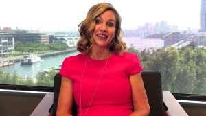 Inman Grant with wavy blonde hair in a bright pink dress and long necklace, seated before a waterfront skyline.
