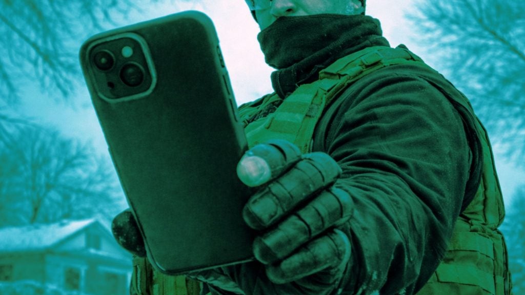 Person wearing tactical vest and gloves with face partially covered, holding a smartphone toward the camera in a snowy yard.