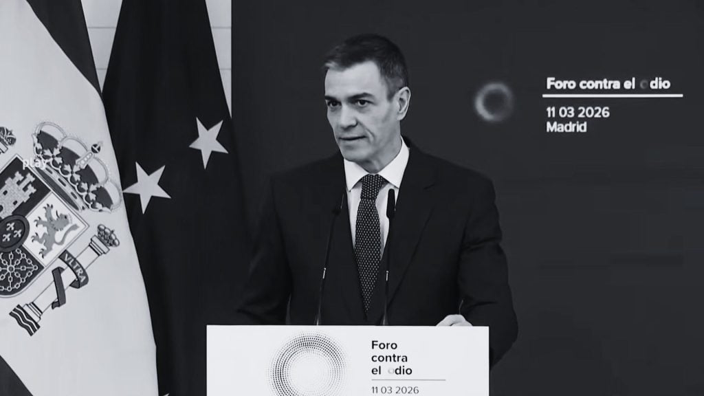 Sanchez in a dark suit and dotted tie speaking at a podium with Spanish flags behind him and text reading "Foro contra el odio 11 03 2026 Madrid."