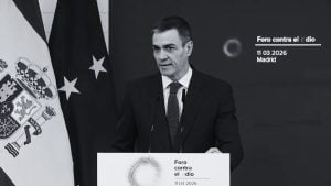 Sanchez in a dark suit and dotted tie speaking at a podium with Spanish flags behind him and text reading "Foro contra el odio 11 03 2026 Madrid."