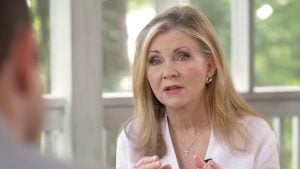 Blackburn with long blonde hair and pearl earrings speaking expressively, wearing a white blouse and cross necklace in a bright room.