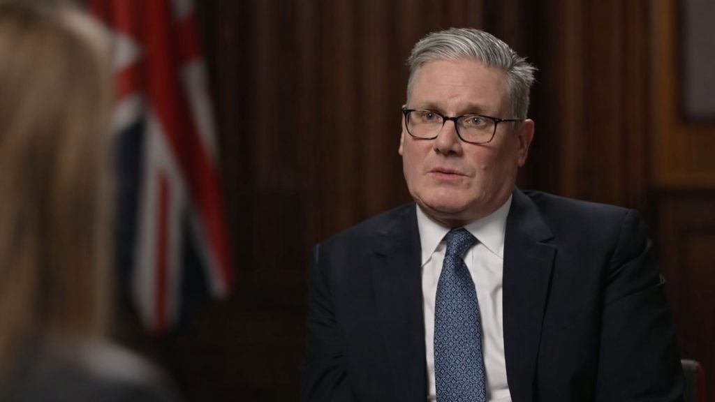 Starmer with gray hair and glasses in a dark suit and patterned tie speaking in a wood-paneled room with a blurred flag behind him.