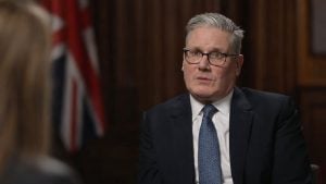 Starmer with gray hair and glasses in a dark suit and patterned tie speaking in a wood-paneled room with a blurred flag behind him.