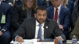 Kash Patel in a dark suit and blue tie speaking into a microphone at a hearing table, papers and water bottles in front of him.