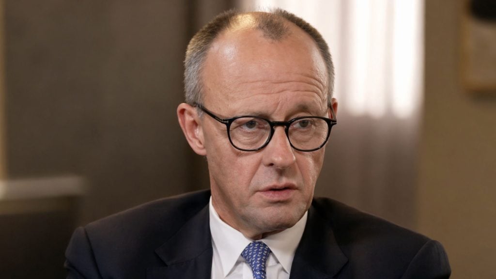 Merz wearing round black glasses, dark suit and patterned blue tie, looking slightly downward in an indoor setting.