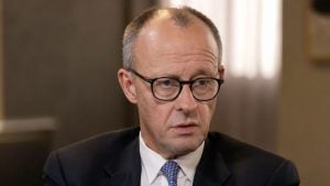 Merz wearing round black glasses, dark suit and patterned blue tie, looking slightly downward in an indoor setting.
